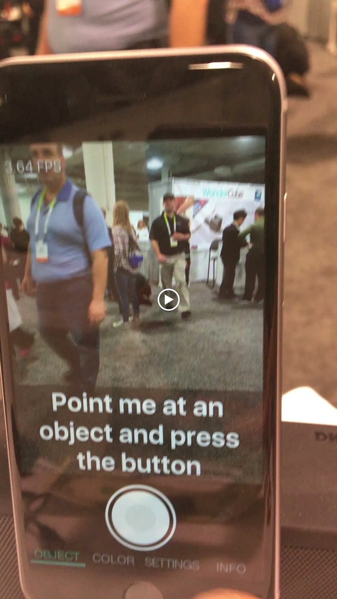 A phone running the Aipoly app on a busy trade-show floor. On screen: ‘Point me at an object and press the button.’