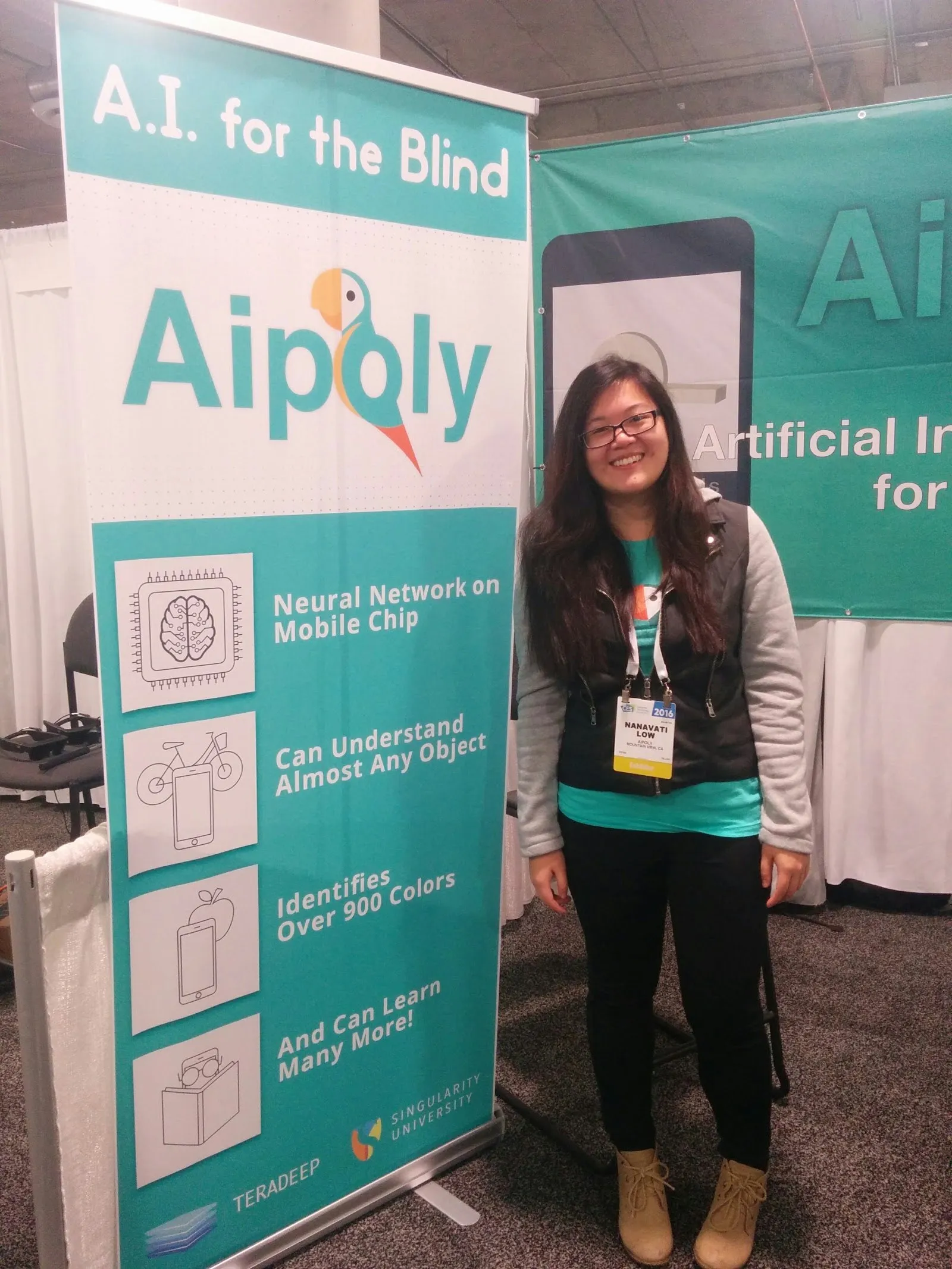 Nanavati Low smiling beside a teal Aipoly banner that reads “A.I. for the Blind”, wearing a CES 2016 badge.